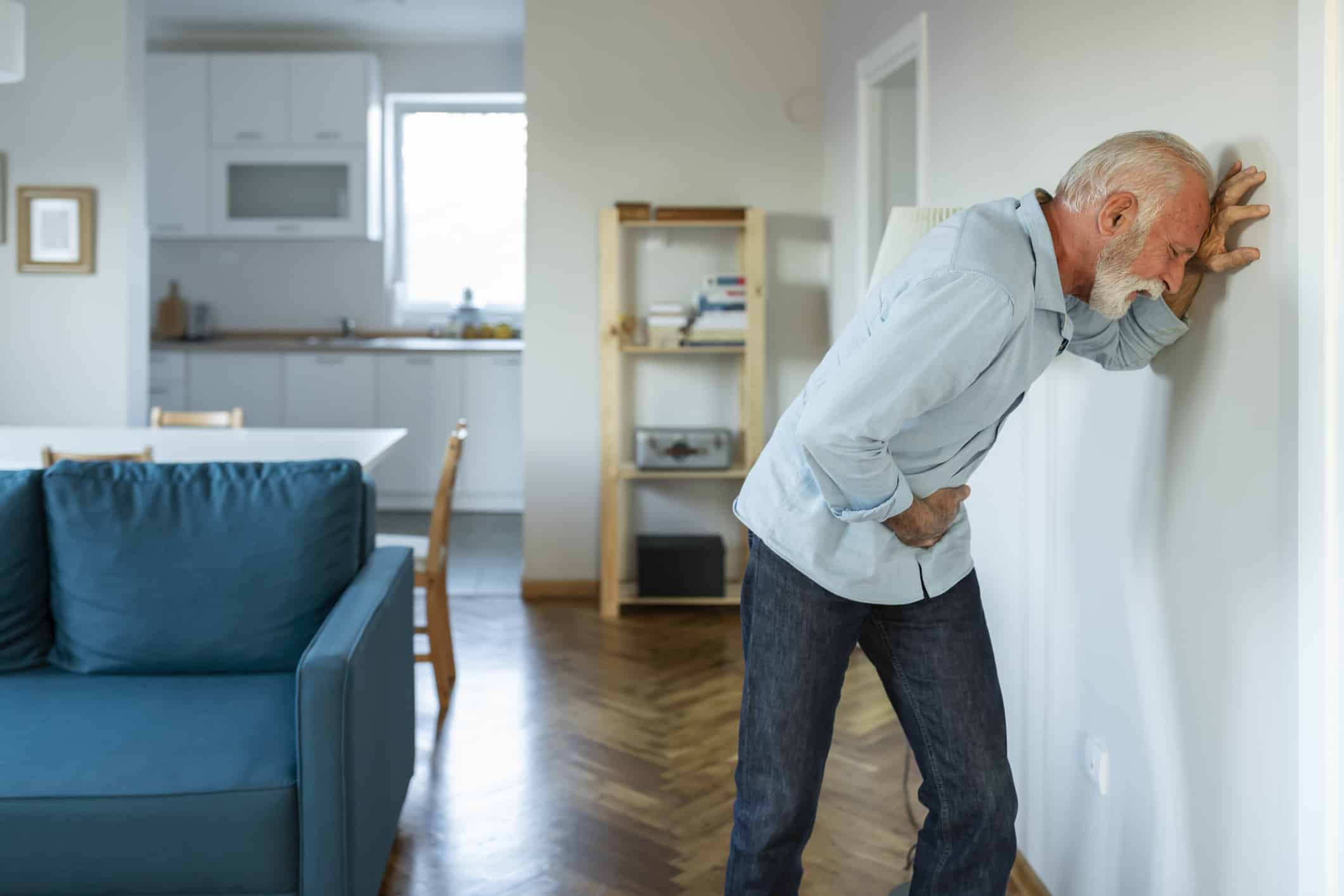 An elderly man leaning on a wall hunched over in his home in serious pain in his pelvis.